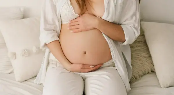 Brzuch ciężarnej opada, gdy zbliża się poród. Kobieta w białej koszuli i biustonoszu delikatnie obejmuje dłońmi swój zaokrąglony brzuch.