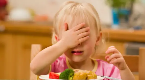 Mała dziewczynka z blond włosami zakrywa oczy ręką, siedząc przy stole z talerzem jedzenia. To obrazek ilustrujący wybiórczość pokarmową u dzieci.