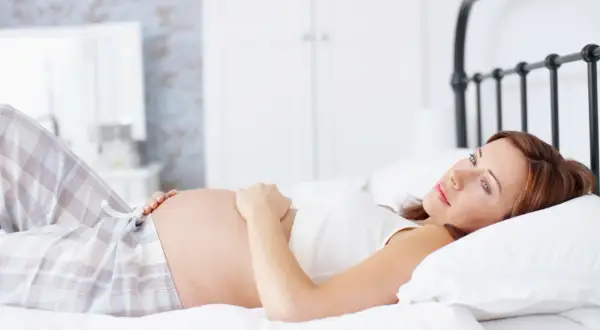 Kobieta w ciąży w pozycji półleżącej na łóżku, delikatnie dotyka brzucha.