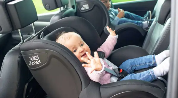 Uśmiechnięte niemowlę w foteliku samochodowym Britax Römer. Obok starsze dziecko. Bezpieczna podróż do jakiego wieku dziecko w foteliku jest kluczowa.