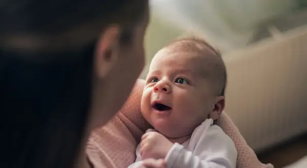 Uśmiechnięty noworodek, trzymany w ramionach, patrzy z zaciekawieniem na mamę. To pierwsze chwile niemowlęcia.