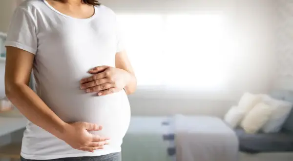 Kobieta w białej koszulce trzyma dłonie na zaokrąglonym brzuchu ciążowym.