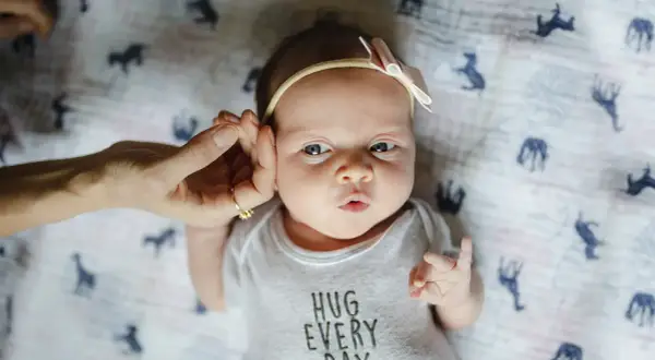 Uroczy, 3-tygodniowe dziecko z kokardką na głowie, w body z napisem "HUG EVERY DAY". Dłoń delikatnie dotyka jego ucha.
