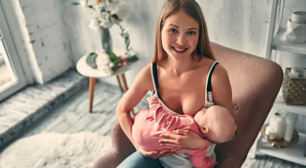 Uśmiechnięta mama karmi piersią dziecko w pozycji klasycznej do karmienia, siedząc w fotelu.