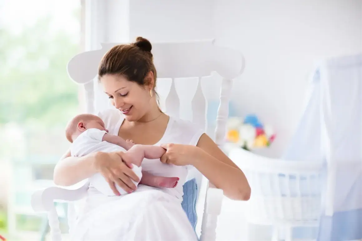 Uśmiechnięta mama kołysze noworodka. W tle widać łóżeczko i kwiaty. To może być moment, gdy zastanawiasz się, jak zatrzymać laktację.