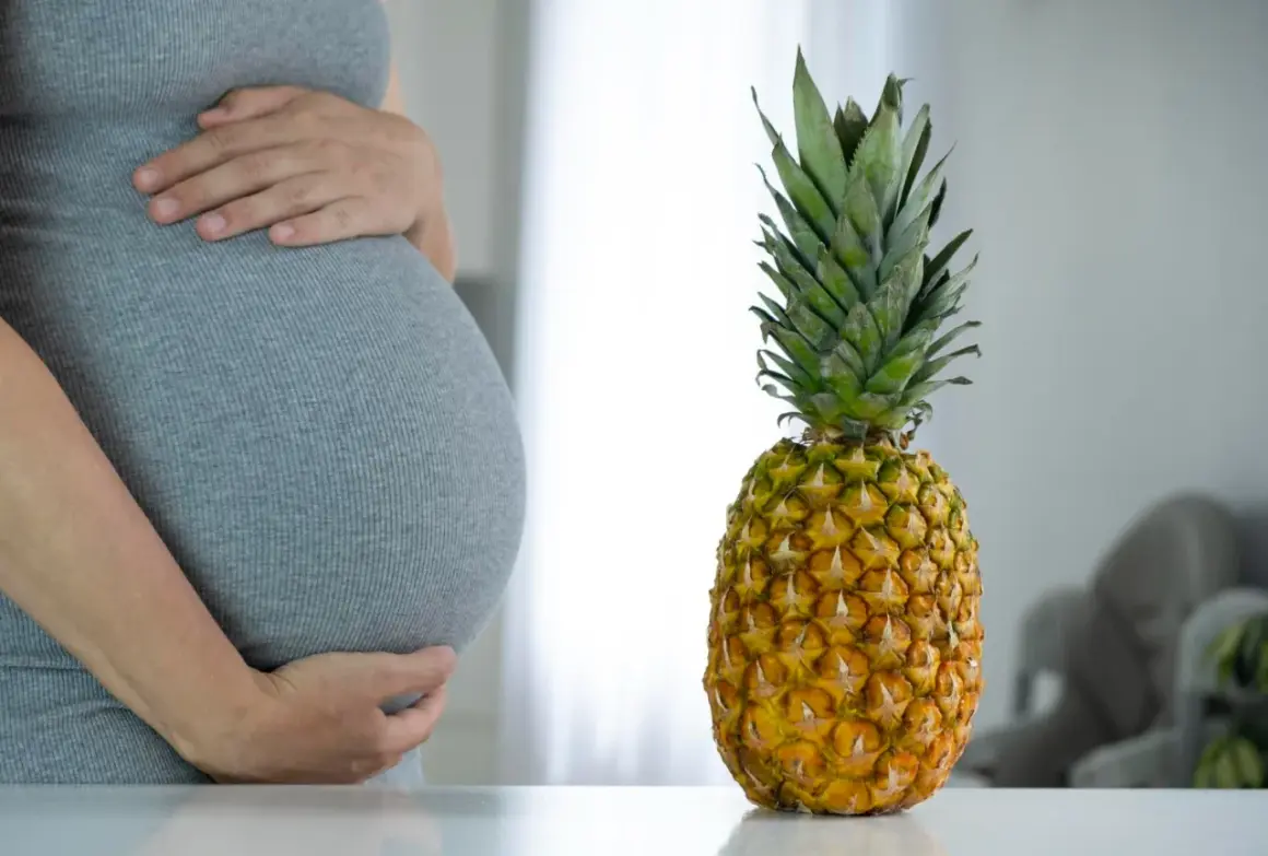 Ciężarna kobieta w szarej sukience trzyma się za brzuch obok ananasa.
