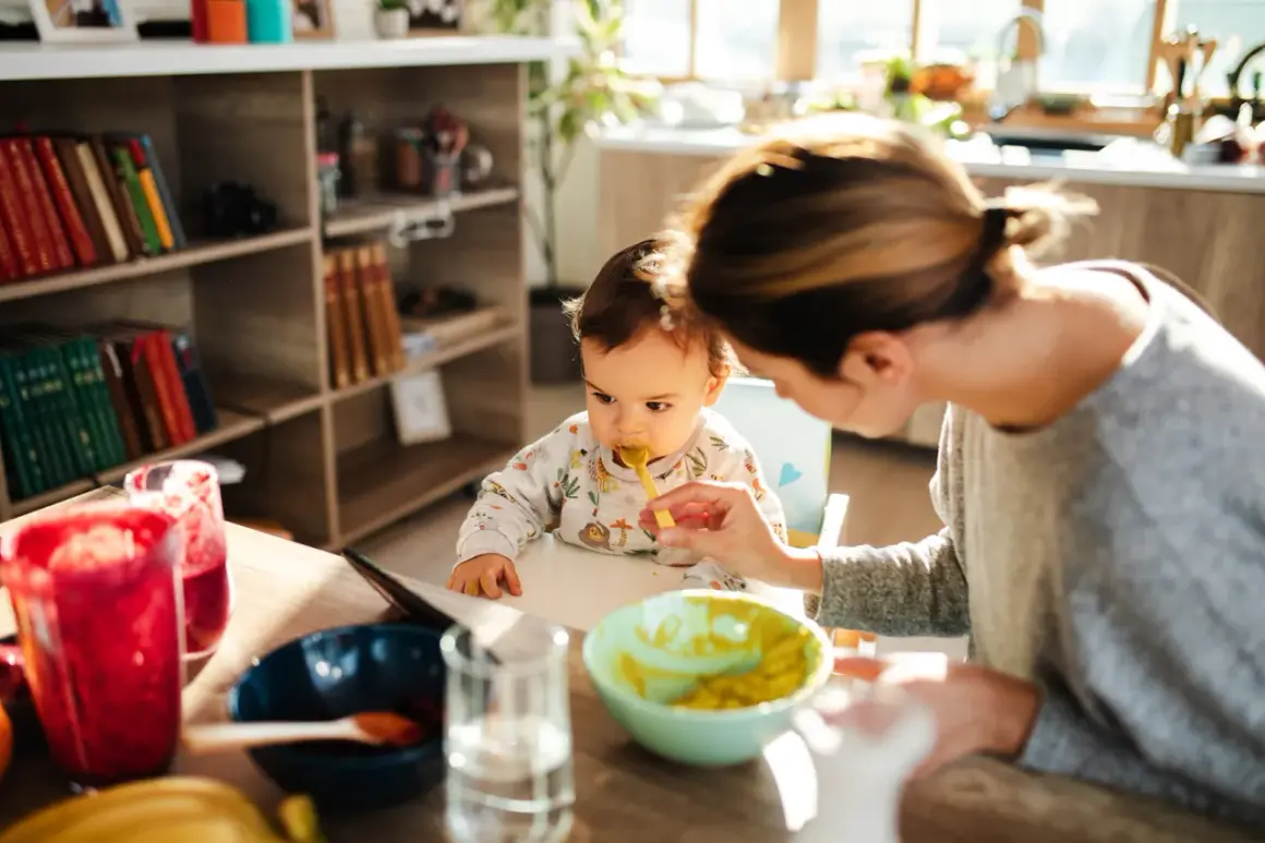Mama karmi dziecko łyżeczką. Na stole są owoce i warzywa, idealne dla niemowlaka, który zaczyna poznawać, co może jeść 6 msc dziecko.