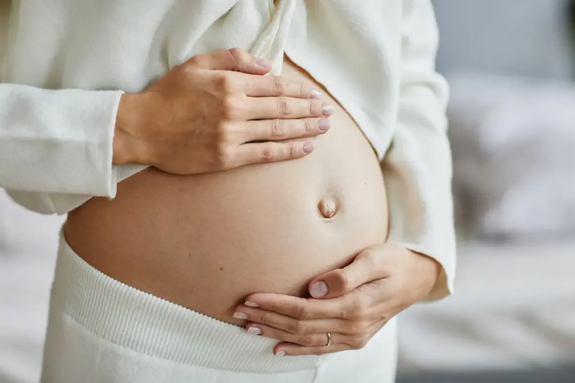Zbliżenie na ciążowy brzuch kobiety, która delikatnie go obejmuje dłońmi.