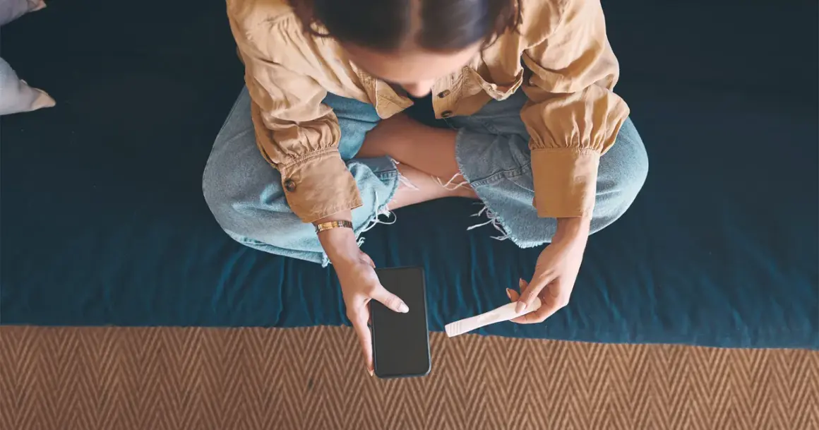 Kobieta siedzi ze skrzyżowanymi nogami, trzymając w dłoniach telefon i test ciążowy.