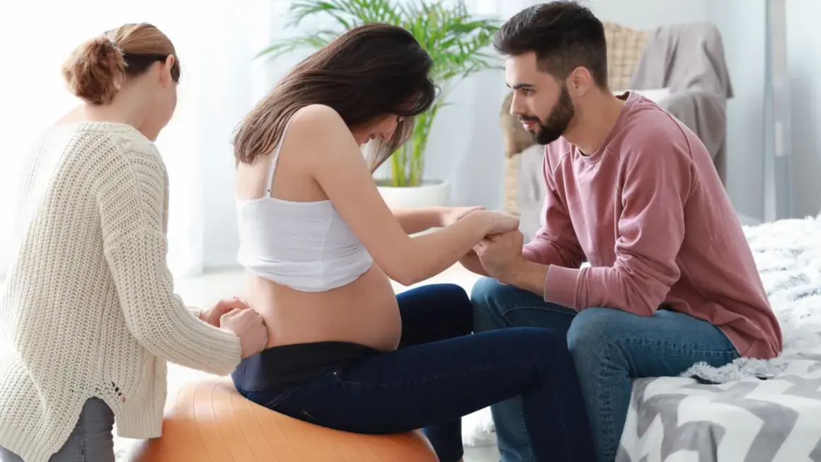 Kobieta w ciąży na piłce, czuć ruchy dziecka. Partner i doula wspierają ją.