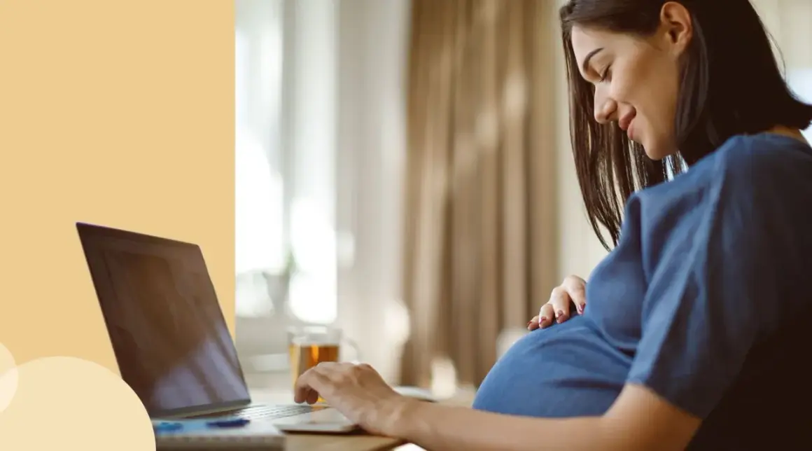 Ciężarna kobieta pracuje przy laptopie, czując ruchy dziecka. Ułożenie główkowe i aktywność malucha są widoczne.