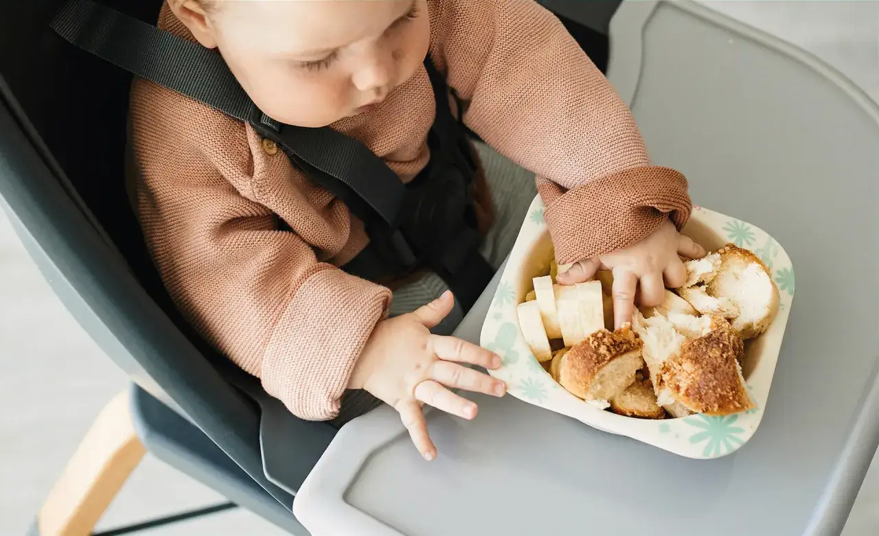 Dziecko w krzesełku próbuje chwycić kawałki chleba i banana. To idealny moment, kiedy rozszerzać dietę dziecka karmionego piersią.