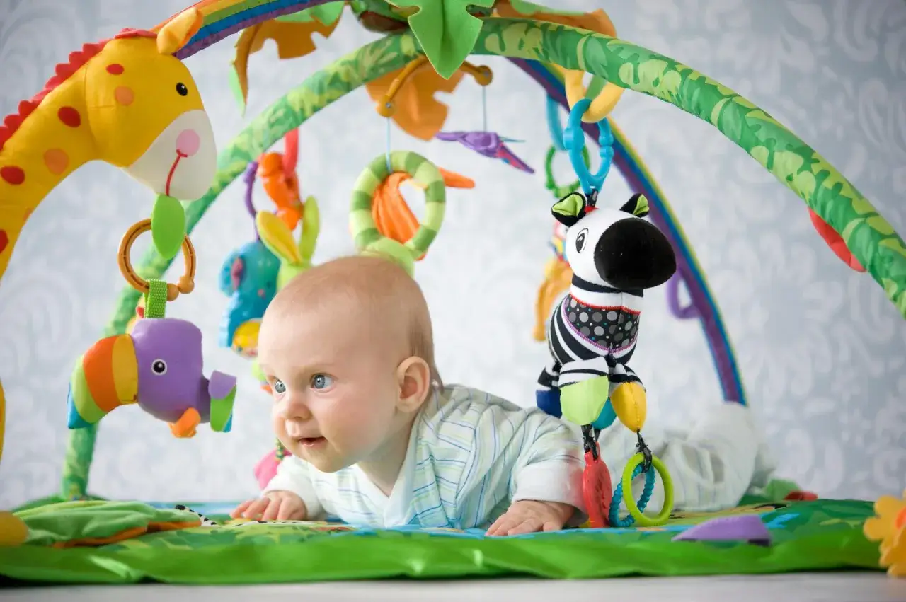 Niemowlę na macie Skip Hop, która jest już wycofana, bawi się wiszącymi zabawkami: żyrafą, tukana i zebrą.