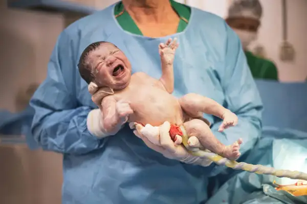 Noworodek z pępowiną, trzymany przez lekarza w rękawiczkach. To moment po porodzie, a nie sposób, jak wywołać poród.