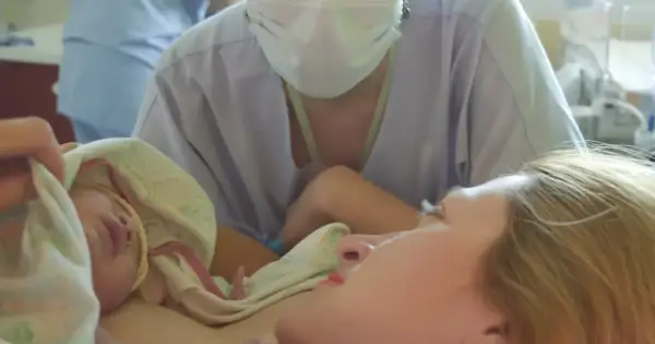 Noworodek przytulony do mamy, otoczony pieluchą. W tle widać personel medyczny w maseczkach, wspierający poród lotosowy.