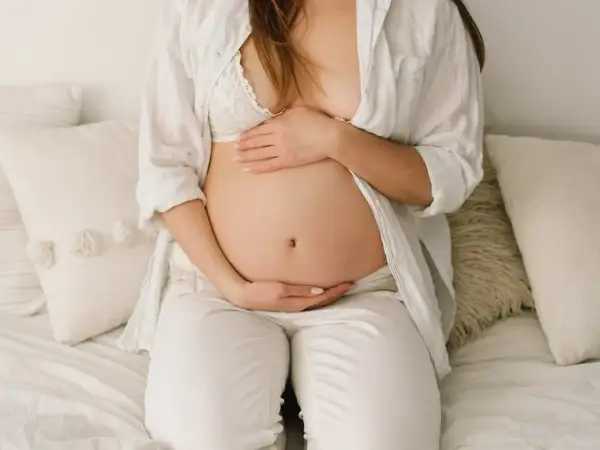 Brzuch ciężarnej opada, gdy zbliża się poród. Kobieta w białej koszuli i biustonoszu delikatnie obejmuje dłońmi swój zaokrąglony brzuch.