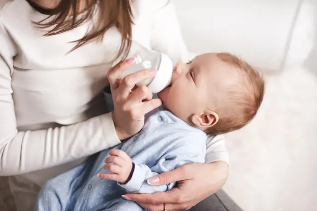 Delikatne karmienie dziecka butelką. Maluch w niebieskim pajacyku pije mleko, czując się bezpiecznie w ramionach mamy.